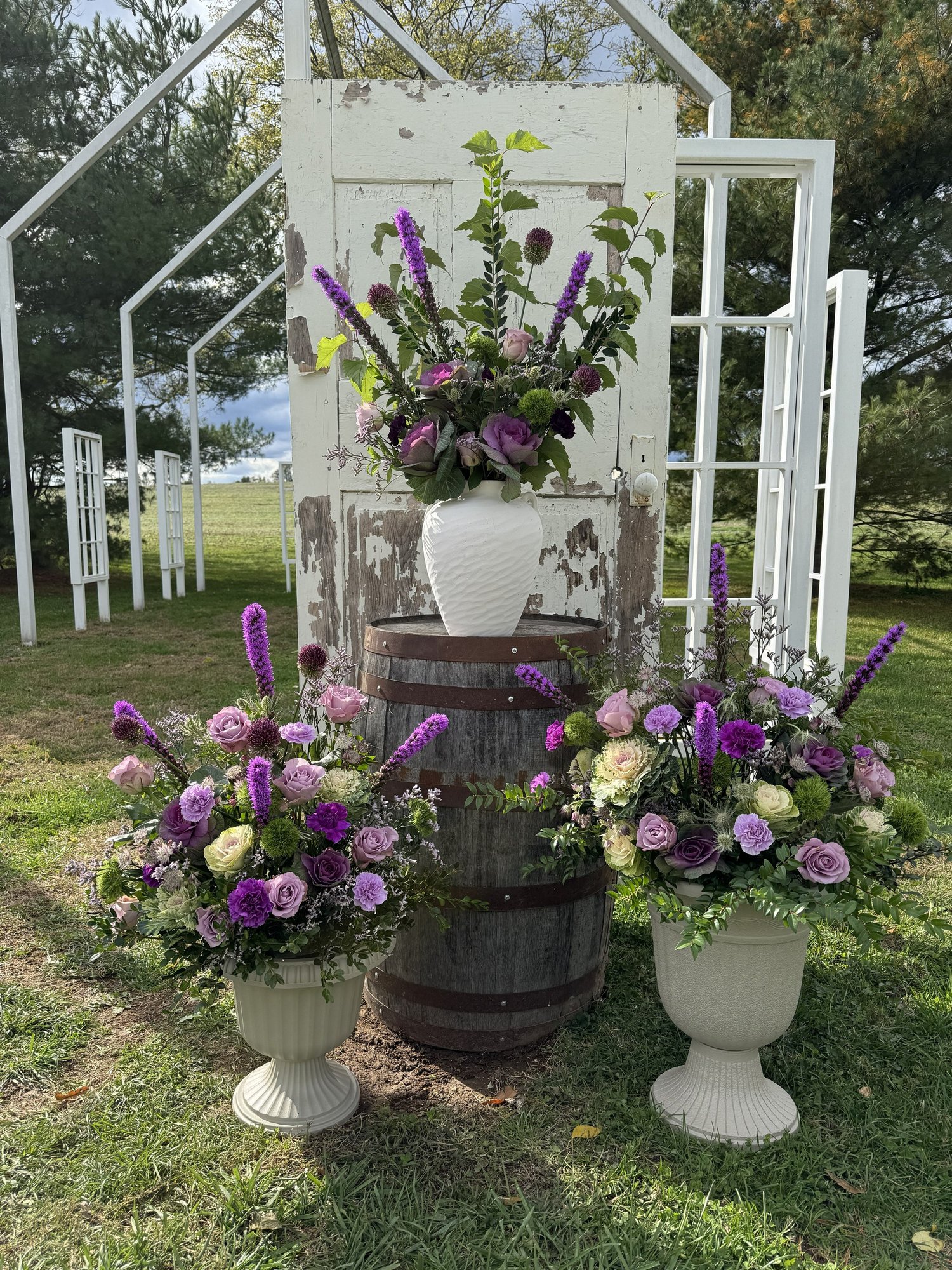 A tall purple arrangement in a white vase on a whiskey barrel, flanked by two urns of roses, kale and liatris