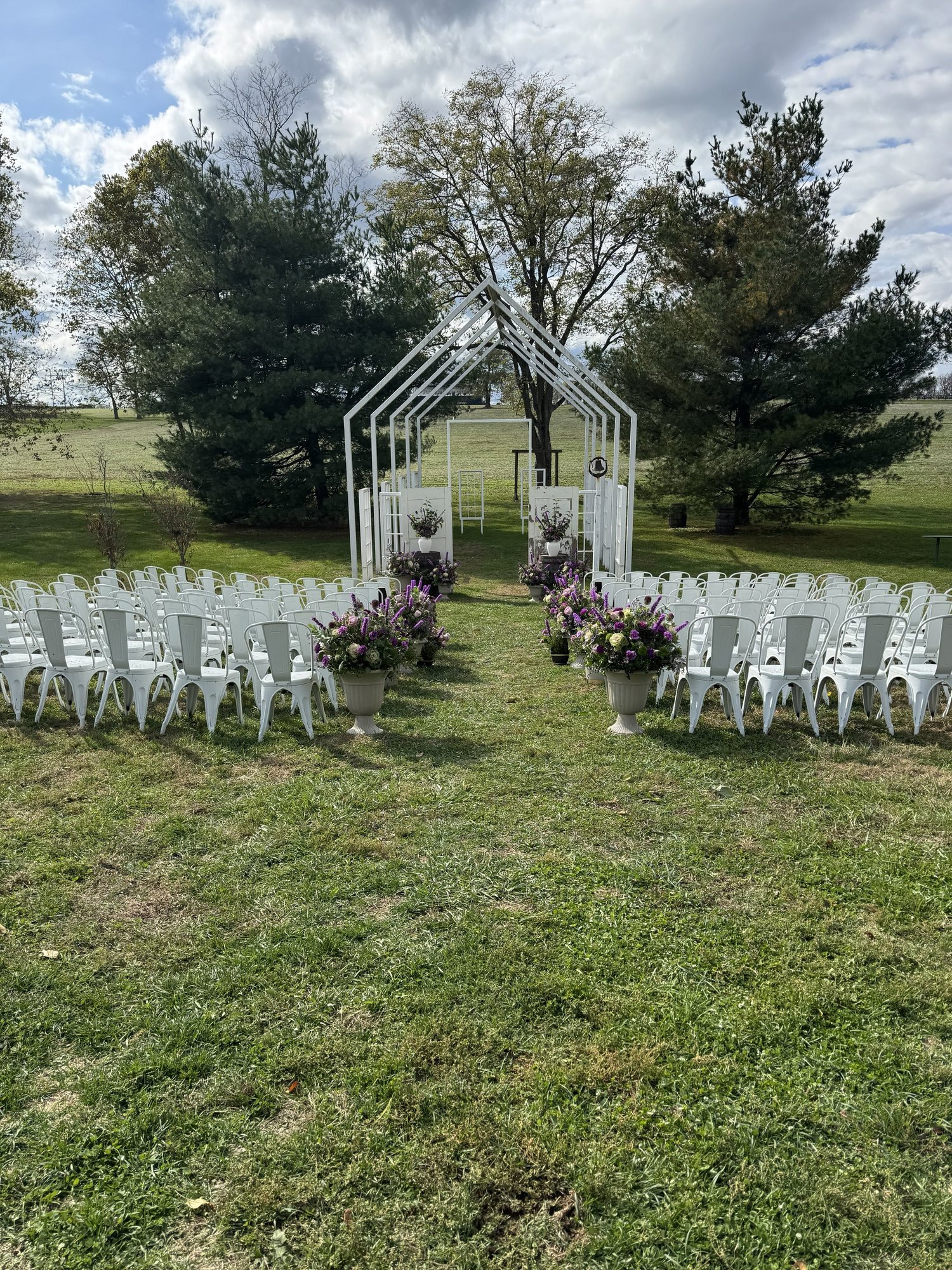 An outdoor ceremony aisle lined with purple and lavender urn arrangements, leading to a white open-frame chapel